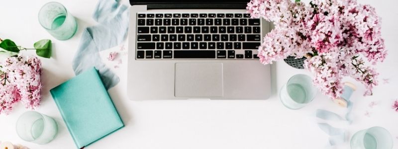 Laptop with blue notebook and pot of pink flowers