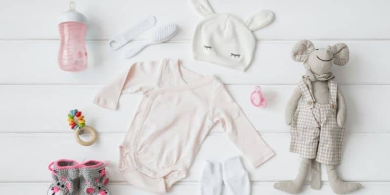 flat lay of baby items on white background, including babygro, hat, brush and comb, pink bottle, pacifier, grey teddy and booties.