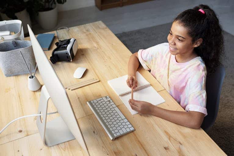 A young girl smiling while studying online at home, using a computer and taking notes.